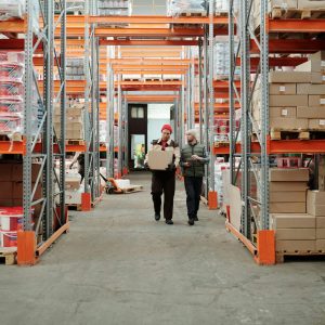 Two workers handle a package in a spacious warehouse surrounded by shelves stocked with boxes and products.